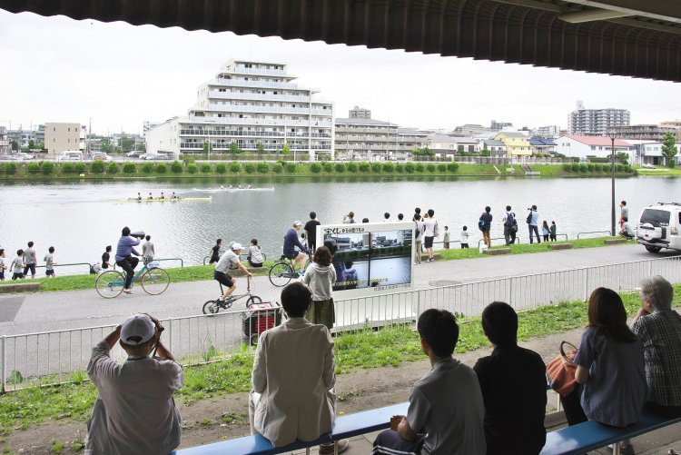 現在の戸田ボートコース。自転車や全速力の応援隊がボートと伴走し、右手の車が実況撮影して観客席のモニターに。