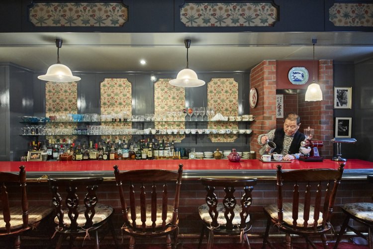 ウィリアム・モリスのひなぎくをあしらった壁紙をバックに『YAMATOYA』店主の熊代さん。