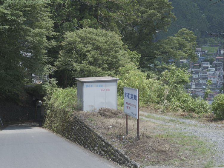 橋梁の先は生活道と同じレベル面になるが敷地には入れない。線路は撤去されている。右手の背後には奥多摩駅停車中のE233系が遠望できる。