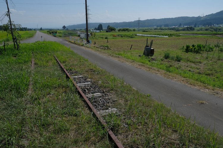 僅かな遺構ではあるけれど、草に埋もれかけたレールが日中線のあった歴史を今に語っている。先は再び生活道路となり、清々しい田園風景が広がっていた。