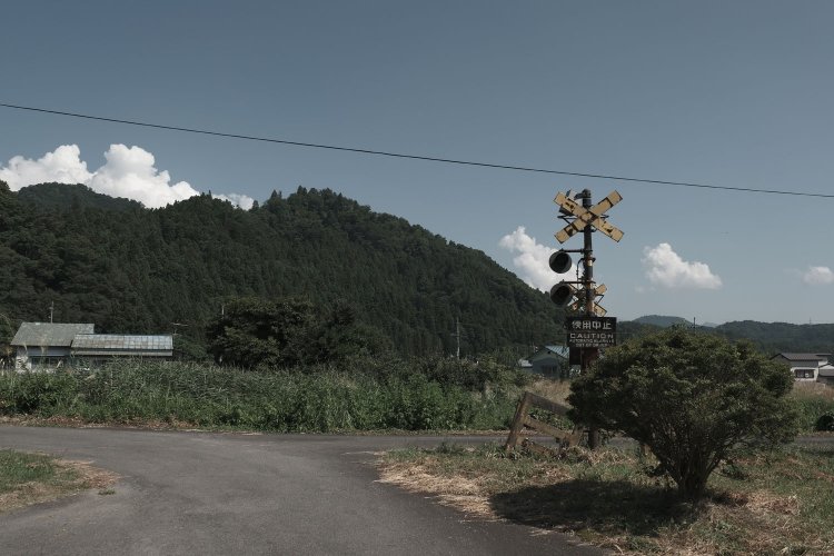 もう二度と鳴らぬ警報器。背後は小山があって家が点々と建つ。夏雲が浮かび、警報器を引き立てる借景となっていた。