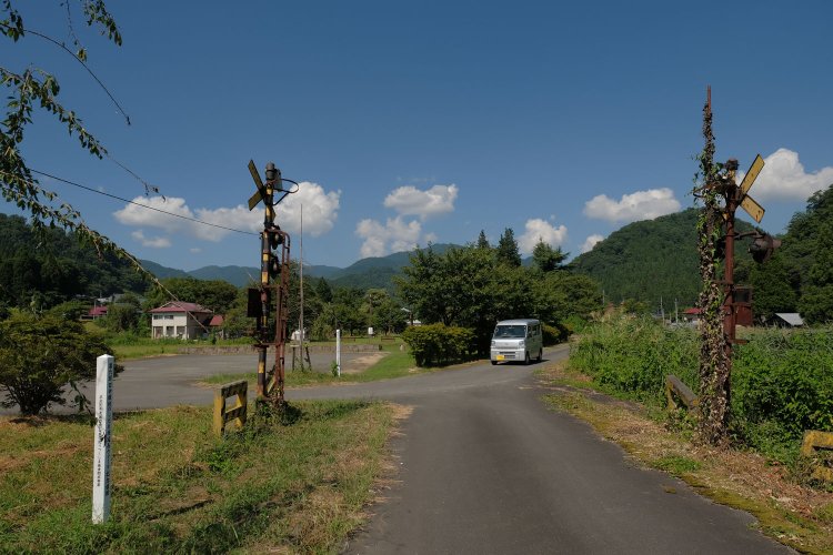 線路であった小道から踏切跡と熱塩駅構内を見る。線路は直線で駅舎へ伸びていたのだろうか。ちょうど軽バンが踏切跡を横切っていく。蔦は警報機を覆い隠すように絡み合う。
