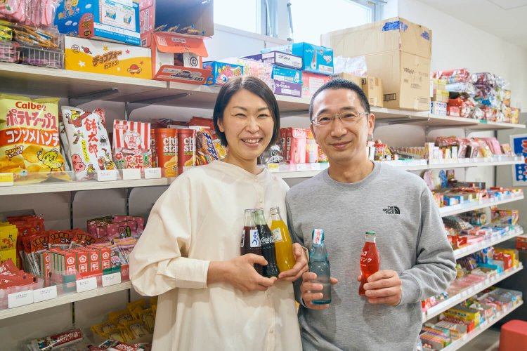 ちびっ子サイダー、柴又ラムネ、瓶ファンタなどを手にする冨田夫妻。駄菓子屋さんらしい品揃え。