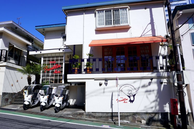 坂の途中に建つ白壁の一軒家。店頭にデリバリーサービスの宅配バイクが並んでいる。
