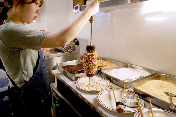 焼き上がったパン生地をアーモンドやココナッツなどでコーティングし、木製トングで棒から外す。