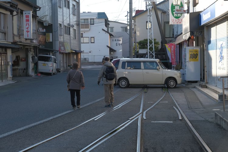 電停の先で軌道が再び集束する。周囲は商店のある町の一角。軌道の先は軽自動車が路駐していた。