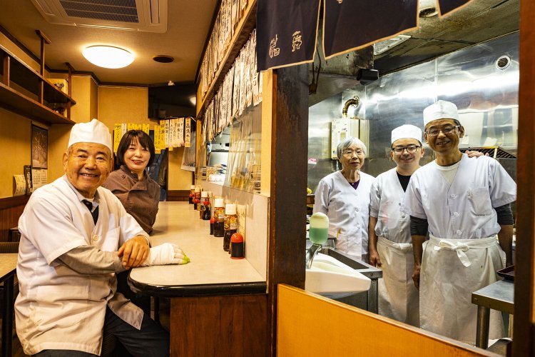 左から2代目の小山芳成さん、女将（3代目妻）の由香さん、厨房内は2代目妻のユキ子さん、4代目の佳祐さん、3代目の昌宏さん。
