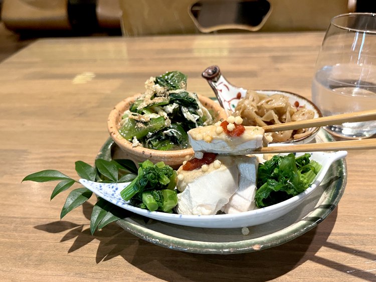 蒸し鶏に酸味のある胡麻みそと豆板醤、香ばしいあられがのっている。両サイドには菜花のお浸しが添えられている。