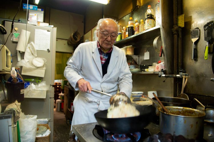 厨房で鍋を振るう武雄さん。一緒に働く娘さんは「夏は冷房もないから心配」と話す。