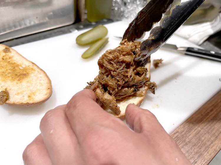 バーベキューソースで味つけた豚肩ロースを焼いてほぐし、それを自家製パンにたっぷり乗せる。さらにピクルス、チェダーチーズを重ねていく。