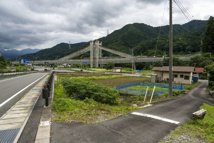 左手が岩島駅方向。旧線跡は左から手前へと線路があり、国道145号によって分断されている。小道の白線部分に踏切の痕跡が残る。奥の立派な橋梁は現在線。
