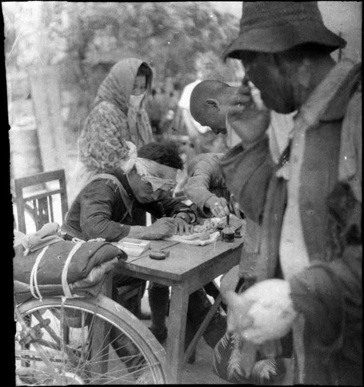 1945年8月6日、撮影=松重美人、中国新聞社所有、日本写真保存センター管理。御幸橋の東側にあった広島地方専売局の前で、被災者に囲まれ「罹災(りさい)証明書」を書く宇品署の藤田徳夫巡査(当時28歳)。