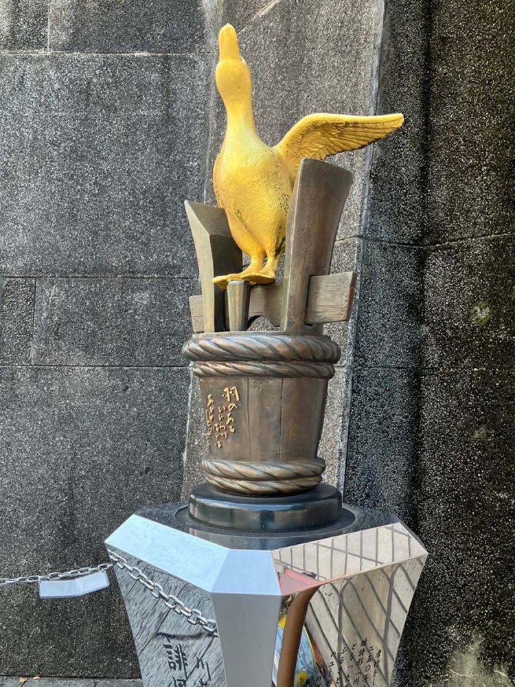 金のあひるが立っている桶部分には、「羽のある　いいわけほどは　あひるとぶ」という川柳が書かれている。