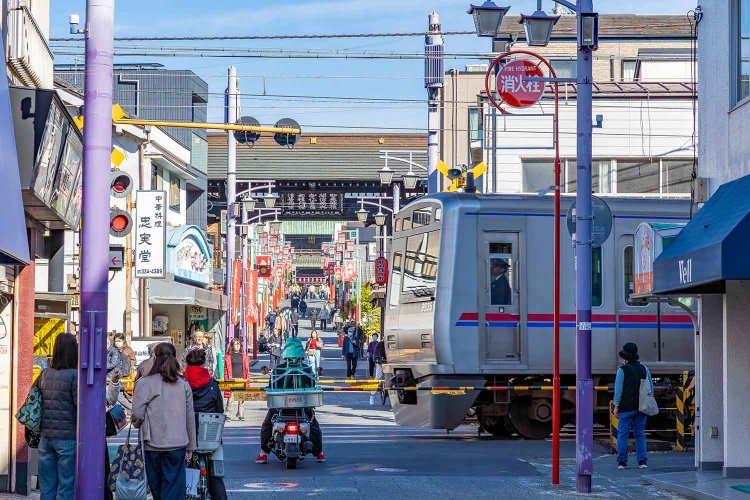 京成本線の踏切とその奥に見える黒門。参道はゆるやかにつづく上り坂だ。