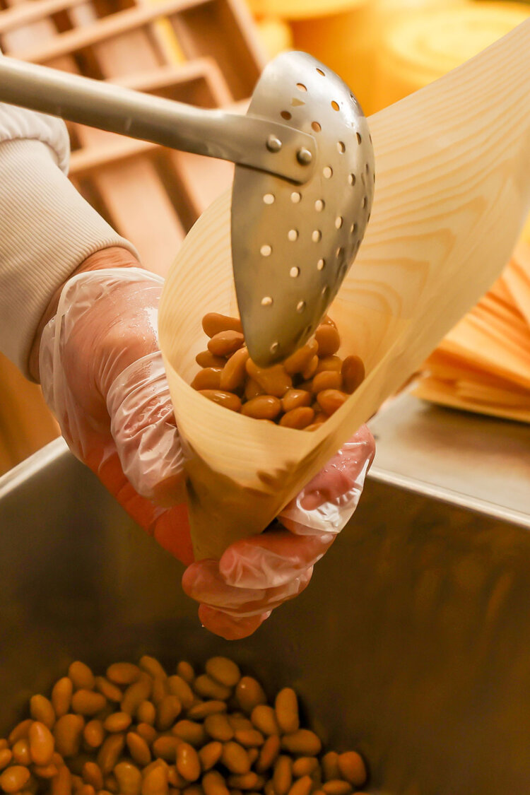 手折りした経木に手詰めする。1パック100g入りで黒豆は80g。