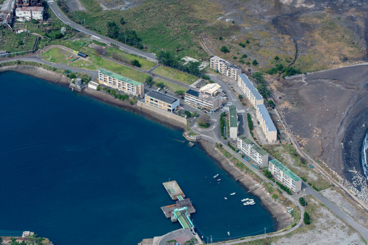 池島港は島の中心地といえる。鉄筋コンクリートのアパートは市営住宅となっている。炭鉱ツアーは港に面した建物が受付だ。写真左上には炭鉱トロッコが見え、道路の真下に廃線跡らしき痕跡も確認できる。