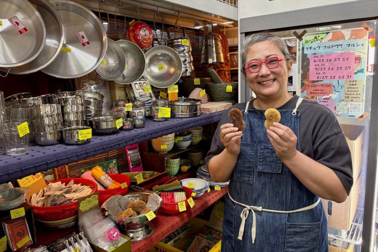 雑貨・食器棚とヨーコさん。手に持つのはスリランカの8の字型ミニたわしだ。